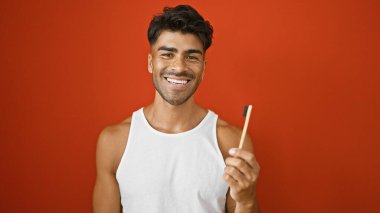 Sakallı, neşeli, genç, İspanyol bir adam, kolsuz bir gömlek içinde kırmızı bir arka plana karşı poz veriyor, elinde kalem var..