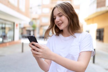 Beyaz bir kadın sokakta akıllı telefon kullanarak gülümsüyor.