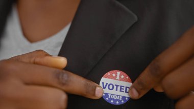 African american woman attaching an 'i voted today' sticker to her blazer in an indoor setting clipart