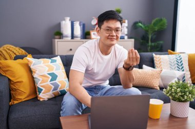 Genç Asyalı adam evinde dizüstü bilgisayar kullanıyor. Kanepede oturuyor. El kol hareketleri yapıyor. 