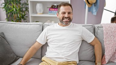 Gri saçlı, ispanyol, olgun bir adam gri bir kanepede rahat bir oturma odasında gülümsüyor..