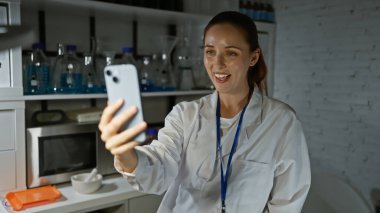 Laboratuvar önlüklü gülümseyen kadın bilim adamı modern laboratuarda selfie çekiyor.