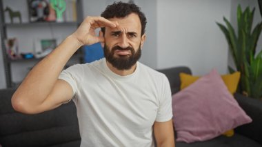 Beyaz tişörtlü sakallı adam arka planda bir bitkiyle evde şaşkın şaşkın bakıyor..