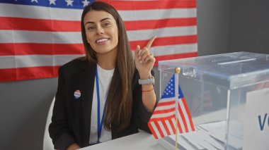 Gülümseyen genç İspanyol kadın oy sandığı ve bayrağıyla Amerikan seçim merkezini işaret ediyor..