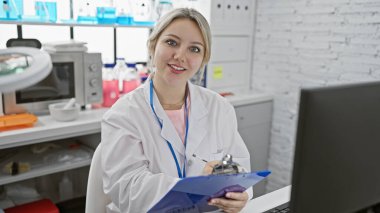 A caucasian woman scientist with blonde hair smiling in a laboratory, holding a clipboard and wearing a lab coat. clipart