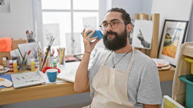 Resim stüdyosunda sakallı yakışıklı bir İspanyol, resim malzemeleriyle çevrili akıllı telefonuyla konuşuyor.