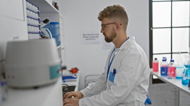 Sakallı, mavi gözlü, laboratuvar önlüğü giyen genç, yakışıklı, beyaz bir adam modern bir laboratuvarda çalışıyor ve bilgisayarda daktilo yazıyor..
