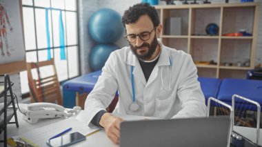 Beyaz laboratuvar önlüklü, orta yaşlı sakallı bir adam bir tıp bürosunda dizüstü bilgisayarda çalışıyor ve profesyonellik yayıyor..