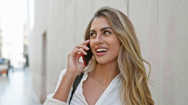 Kendine güvenen, mutlu, genç sarışın kadın akıllı telefonuyla konuşmaktan zevk alıyor, güneşli, şehir sokağında canlı bir ifadeyle dikiliyor..