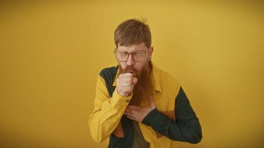 Kızıl saçlı, gözlüklü, sarı arka planda öksüren bir adam..