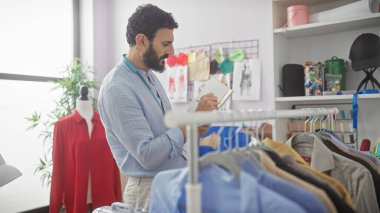 Boynundaki bantla yakışıklı sakallı terzi, renkli giysilerle çevrili, güneşli bir atölyede tasarımlar yapıyor..
