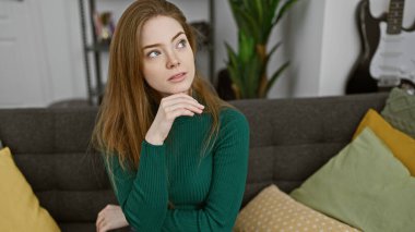 Düşünceli genç bir kadın rahat bir oturma odasında oturur. Arka planda minderleri ve gitarı vardır..