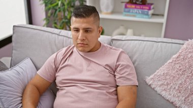 Oturma odasındaki kanepede ciddi bir ifadeyle oturan genç latin adam, potansiyel olarak endişeli ya da derin düşüncelere dalmış, kapalı alandaki sorunları düşünüyor.