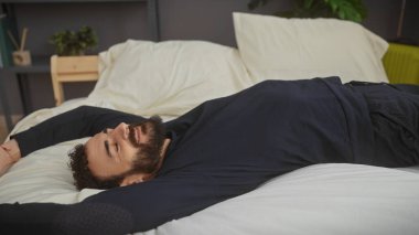 Sakallı yakışıklı bir adam, rahat bir yatak odasındaki bir yatakta dinleniyor. Rahatlama ve ev içi rahatlığı çağrıştırıyor..