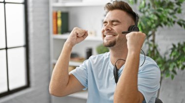 Sakallı, neşeli, genç, İspanyol bir adam modern bir iç mekan ortamında kulaklık takarak başarıyı kutluyor..