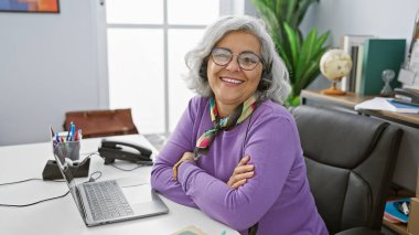 Gri saçlı, gözlüklü ve modern ofis ortamında kulaklık takan gülümseyen olgun bir kadın..