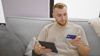 Genç sakallı bir adam evinde gri bir kanepeye oturmuş elinde bir tablet ve kredi kartı tutuyor..
