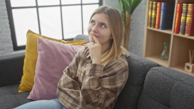 Düşünceli genç bir kadın, renkli yastıklar ve kitaplıklarla çevrili, rahat bir oturma odasında, gri bir kanepede oturuyor..