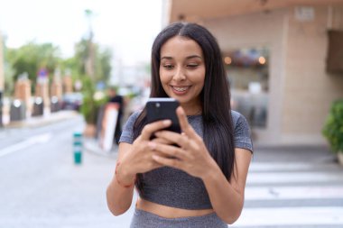 Genç Arap kadın sokakta akıllı telefon kullanarak kendine güvenerek gülümsüyor.