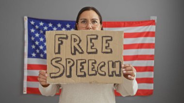Hispanic woman holding a free speech sign in front of an american flag indoors. clipart