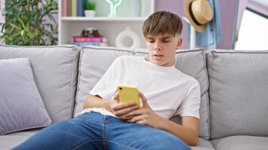 Caucasian teenage boy lounging on sofa at home while scrolling through his smartphone clipart