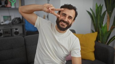 Gülümseyen sakallı adam, modern bir oturma odasında bitkilerle barış işareti yapıyor.
