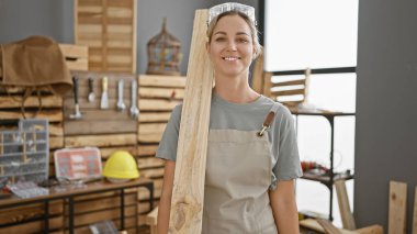 A cheerful young woman with blonde hair, wearing safety glasses and an apron, holds lumber at a carpentry workshop indoors. clipart