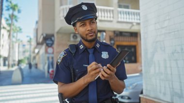Afro-Amerikan polisi güneşli bir şehir manzarasında bir tablete not alıyor.