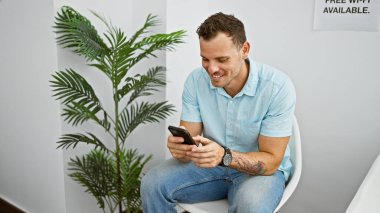 Dövmeli, gülümseyen İspanyol adam modern bekleme odasında akıllı telefon kullanıyor, gelişigüzel ve birbirine bağlı bir atmosfer yaratıyor..
