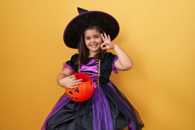 Cadı kostümü içinde sırıtan neşeli İspanyol kız, elinde balkabağı sepeti, parmaklarıyla kendine güvenen bir 