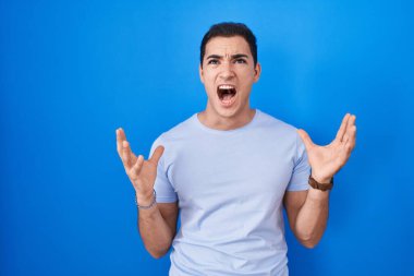 Mavi arka planda dikilen Latin asıllı genç adam kızgın ve deli gibi bağırıyor ve agresif bir ifade ve kollarla bağırıyor. hüsran kavramı. 