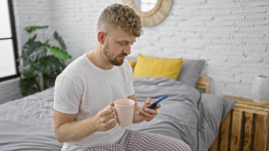Akıllı telefon kullanarak ve çağdaş bir yatak odasında kahve fincanı tutarak günlük giysiler içinde sakallı yakışıklı bir genç.