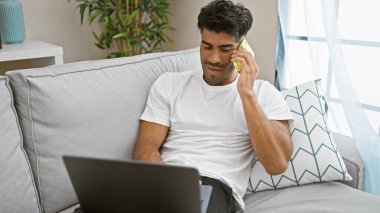 Genç, yakışıklı, İspanyol bir adam telefonda dizüstü bilgisayar kullanarak konuşuyor. Modern bir oturma odasındaki kanepede oturuyor..