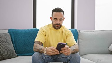 Odaklanmış, sakallı genç bir İspanyol adam akıllı telefonunu kullanarak evde oturuyor ve sıradan bir ev hayatı sahnesini canlandırıyor..