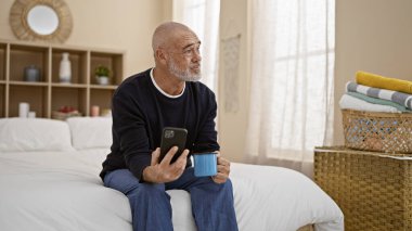Serene orta yaşlı, gri sakallı, elinde akıllı telefon ve kahve fincanı olan bir adam.