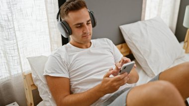 Beyaz tişörtlü yakışıklı genç adam rahat bir yatak odasında kulaklık ve akıllı telefonla rahatlıyor.