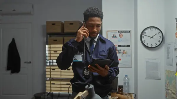 stock image African american detective multitasking in a police station, examining a tablet while talking on the phone indoors.