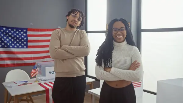 stock image A man and a woman with arms crossed stand smiling in an indoor american electoral setting.