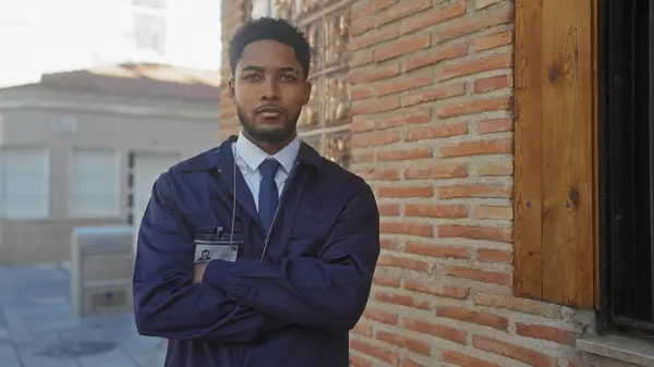 stock image Confident african american man, wearing a formal blue uniform, stands with arms crossed on a city street, exuding professionalism and authority.