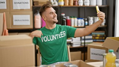 Gülümseyen İspanyol adam bağış kutularıyla dolu bir depoda yeşil gönüllü tişörtü giyerek selfie çekiyor..