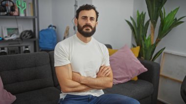 Beyaz gömlekli, orta yaşlı, ciddi bir sakallı adam modern bir oturma odasında kollarını kavuşturmuş oturuyor..