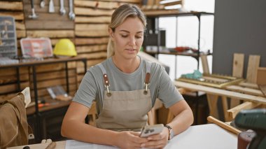 Atölyedeki sarışın kadın arka planda aletleri ve tahtası olan akıllı telefon kullanıyor.