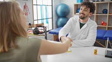 Bir erkek doktor ve kadın hasta, kliniğin fizyoterapi odasında rehabilitasyon ekipmanlarıyla el sıkışıyor..