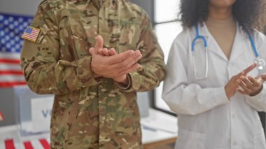 Askeri üniformalı bir adam ve tıbbi giyinmiş bir kadın arka planda ABD bayrağıyla içeride alkışlıyorlar..