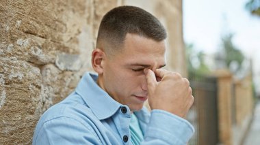 Günlük giysiler içinde stresli bir İspanyol genç şehir ortamında duruyor, burnunun köprüsüne masaj yapıyor..