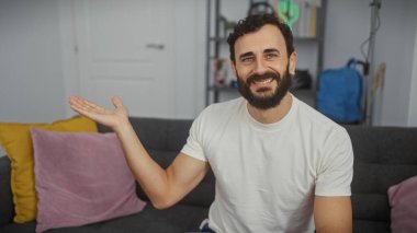 Beyaz tişörtlü, gülümseyen sakallı adam modern oturma odasında bir şey sunuyor..