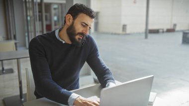 Modern ofis ortamında dizüstü bilgisayar kullanan sakallı adam profesyonelliği ve konsantrasyonu yansıtıyor..