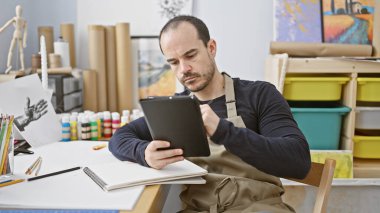 Stüdyoda sakallı İspanyol bir adam sanat referansları için tablet kullanıyor. Etrafı yaratıcı aletler ve tuvallerle çevrili..