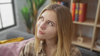 Düşünceli, genç bir kadın, sarı saçlı rahat bir oturma odasına bakıyor..