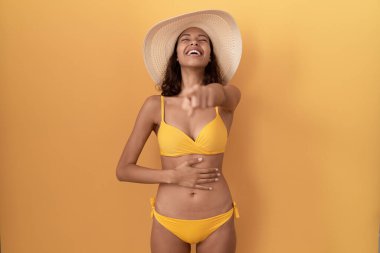 Bikini giyen ve yaz şapkası takan genç İspanyol kadın sana gülüyor, eli vücudunun üzerinde kamerayı işaret ediyor, utanç ifadesi. 